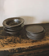 Ceramic Bowl i Black Glazed , en bolle, fra 10AM Apoteke, et livsstilsmarkedet fra 10AM Loft. Produsert i kearmikk, sort leire og håndlaget i Hellas. Kollekted by en intedriørforretning på Grünerløkka i Oslo forhandler av 10AM Apoteke. Bildet viser 4 boller og 7 tallerkener i et varmt gresk hjem